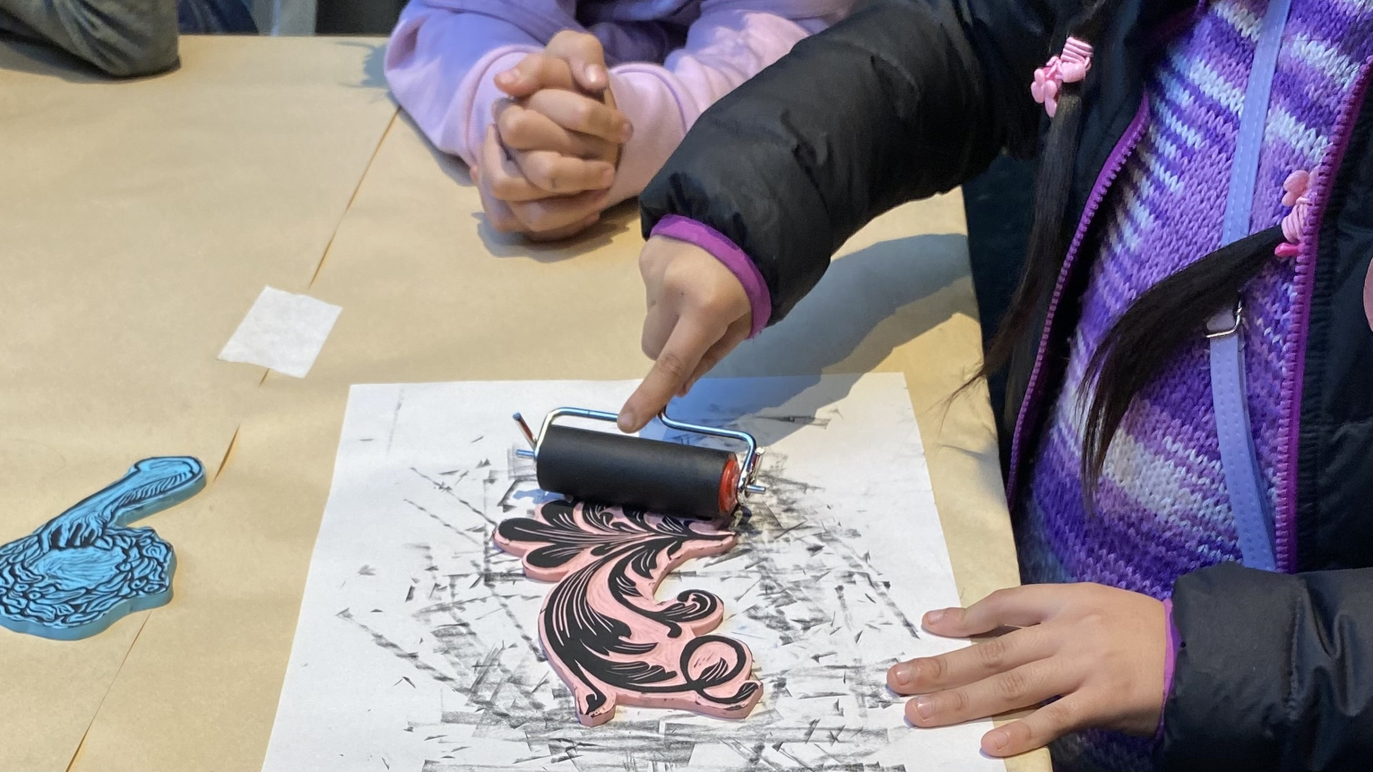 Fotografía de las manos de una niña imprimiendo una figura sobre una hoja de papel, con un rodillo con tinta