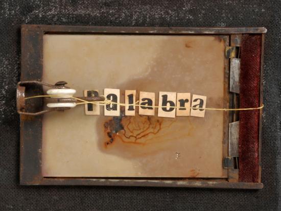 Fotografía de una especie de antigua trampa para ratones, con trozos de papel  en los que se deletrea "palabra"
