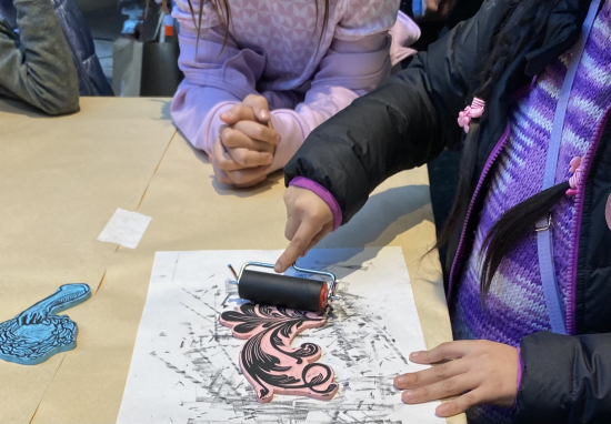 Fotografía de las manos de una niña imprimiendo una figura sobre una hoja de papel, con un rodillo con tinta